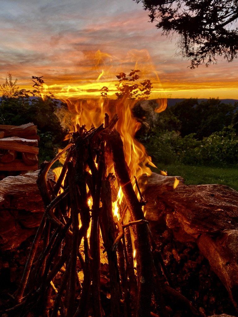 A campfire burns with bright orange flames against a backdrop of a colorful, sunset-lit sky and distant hills.