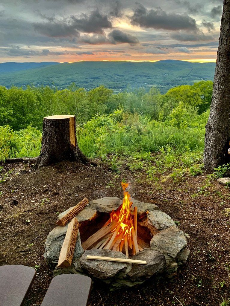 A fire crackling in a stone fire pit overlooks a lush, mountainous landscape under a cloudy sunset sky.