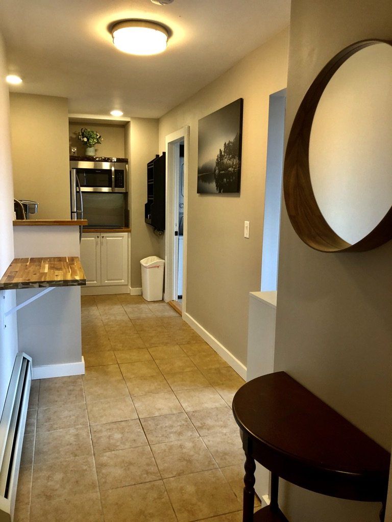 An interior view of a hallway leading to a kitchen with beige tiled floors, a round mirror, and a small console table.