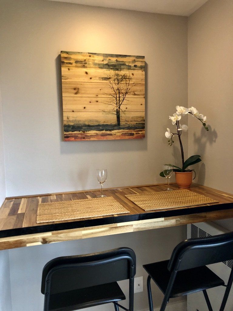 A wood-plank wall art piece hangs above a table with two black chairs, place mats, and a white orchid in a pot.