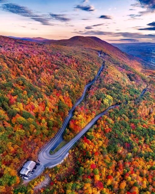 An aerial view of a winding mountain road through a dense, colorful autumn forest at sunset.