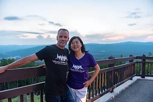 A couple wearing matching shirts stands on an observation deck overlooking a mountainous landscape at twilight.