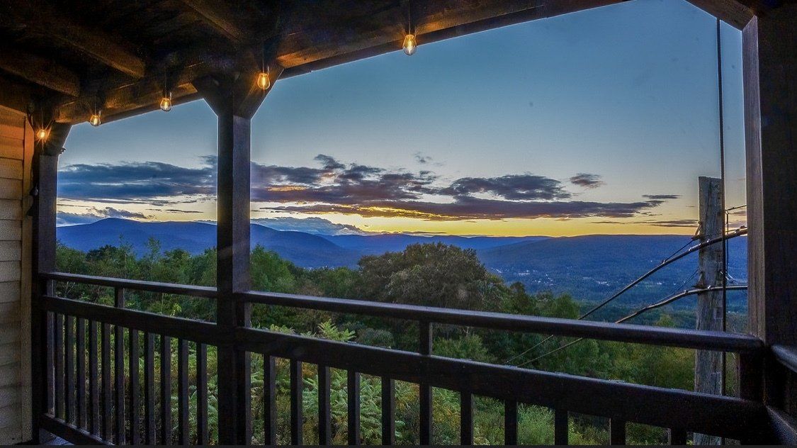 A wooden balcony overlooks a forested valley under a twilight sky with soft, colorful clouds at sunset.