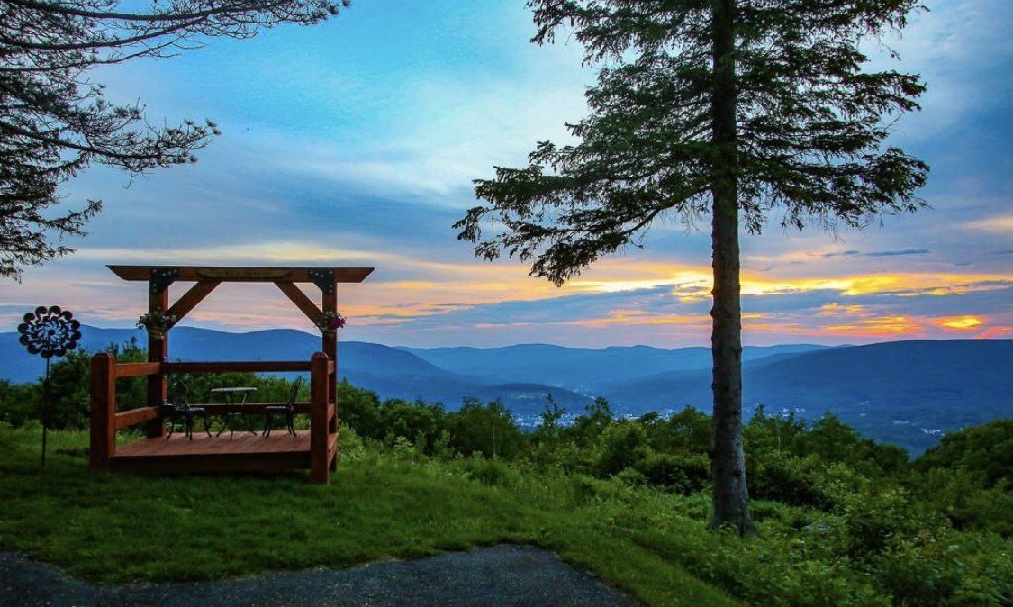 A wooden pergola overlooks a scenic mountain valley at sunset, with a large tree in the foreground.