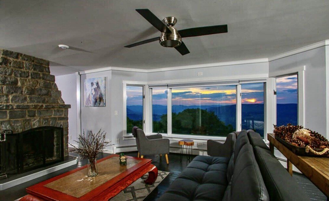 Living room with a stone fireplace, red coffee table, grey sofa, and large windows revealing a scenic mountain sunset.