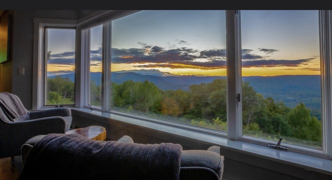 A view from a cozy room with comfortable seating through a large window overlooking a mountain sunset and forest.