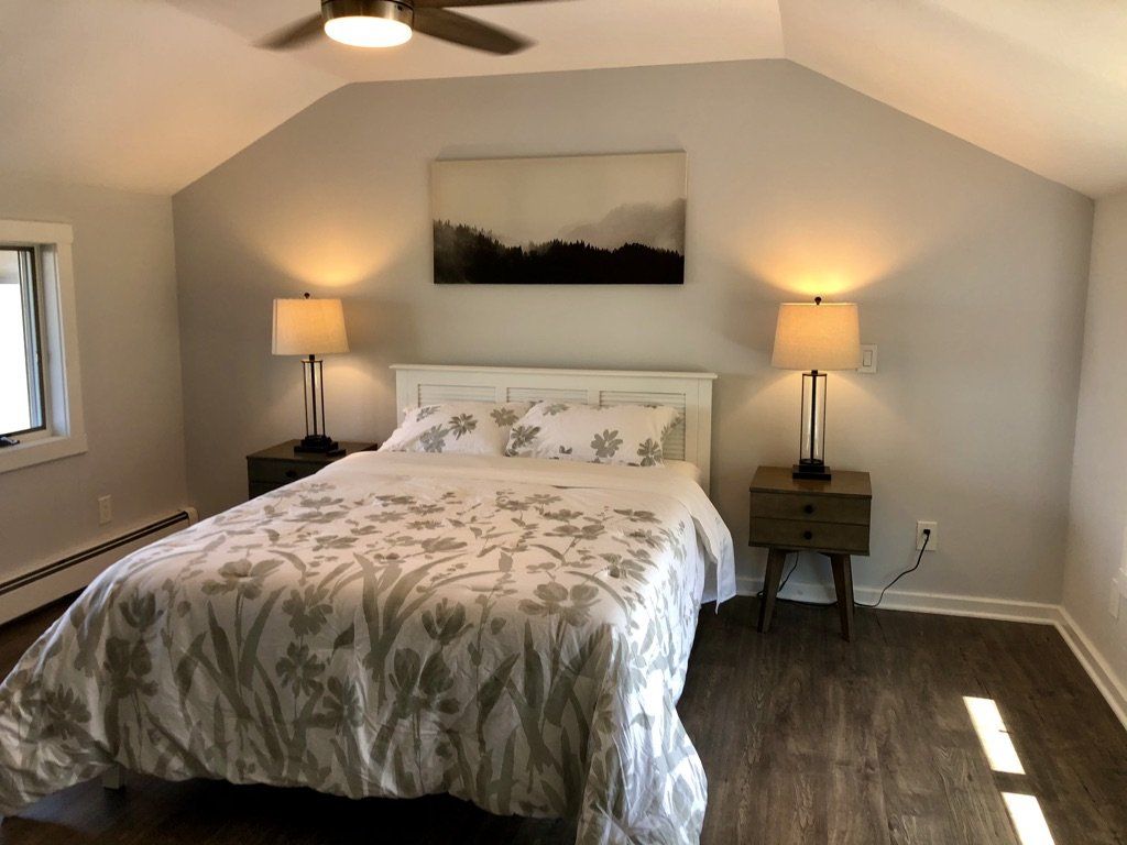 A cozy bedroom featuring a queen bed with white floral bedding, dual nightstands with lamps, and a landscape wall art.