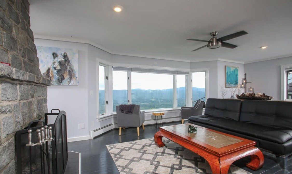 A modern living room with a stone fireplace, black sofa, wooden coffee table, and large bay windows overlooking mountains.