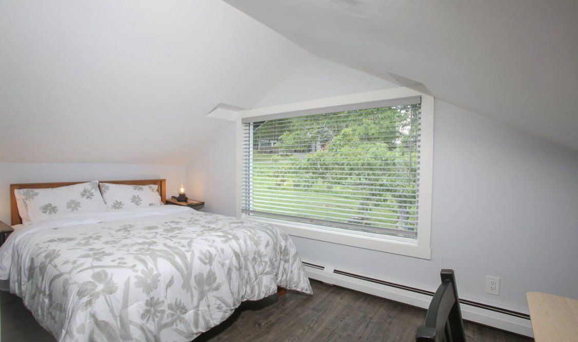 A bedroom with a sloped ceiling, featuring a bed with white patterned bedding and a window looking out onto greenery.