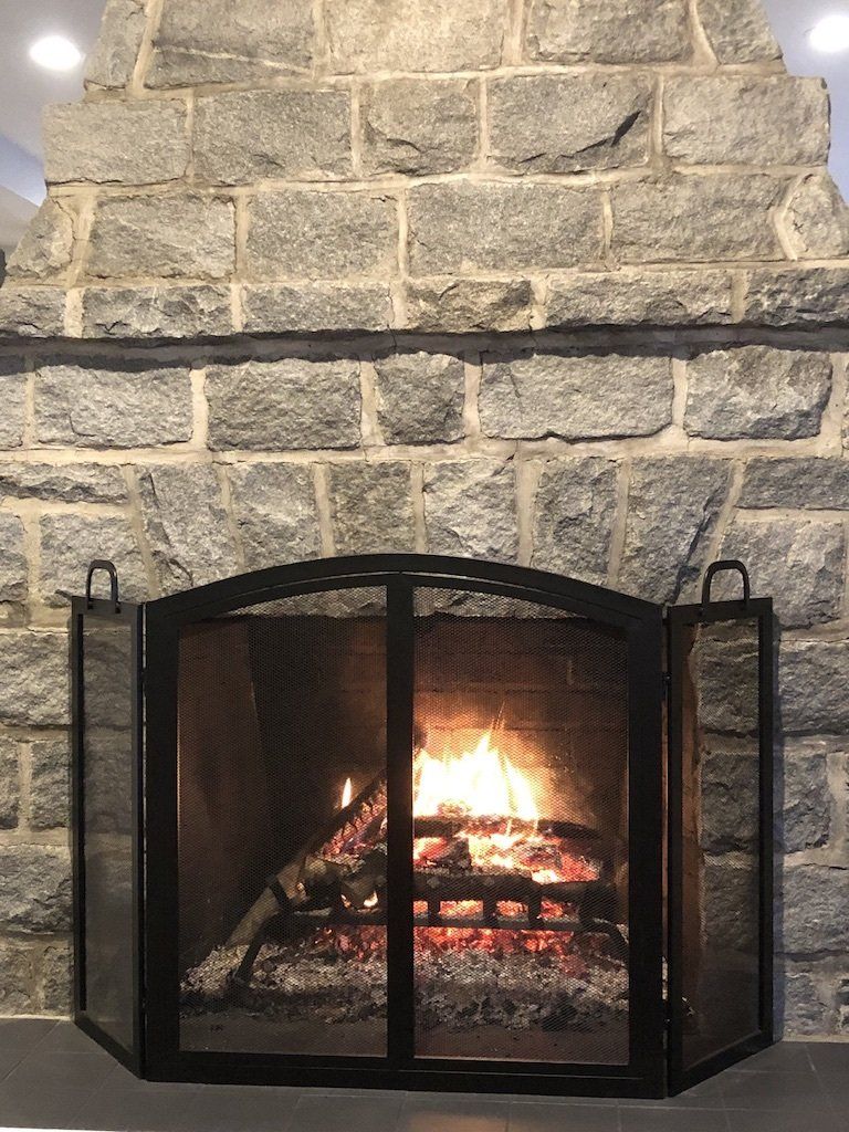 A burning fire inside a stone fireplace, protected by a black, three-panel metal mesh screen.