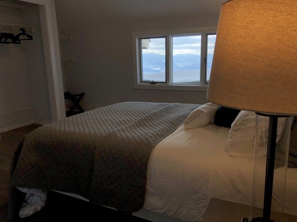 A bedroom with a bed featuring a gray patterned duvet and white pillows, illuminated by a lamp in the foreground.