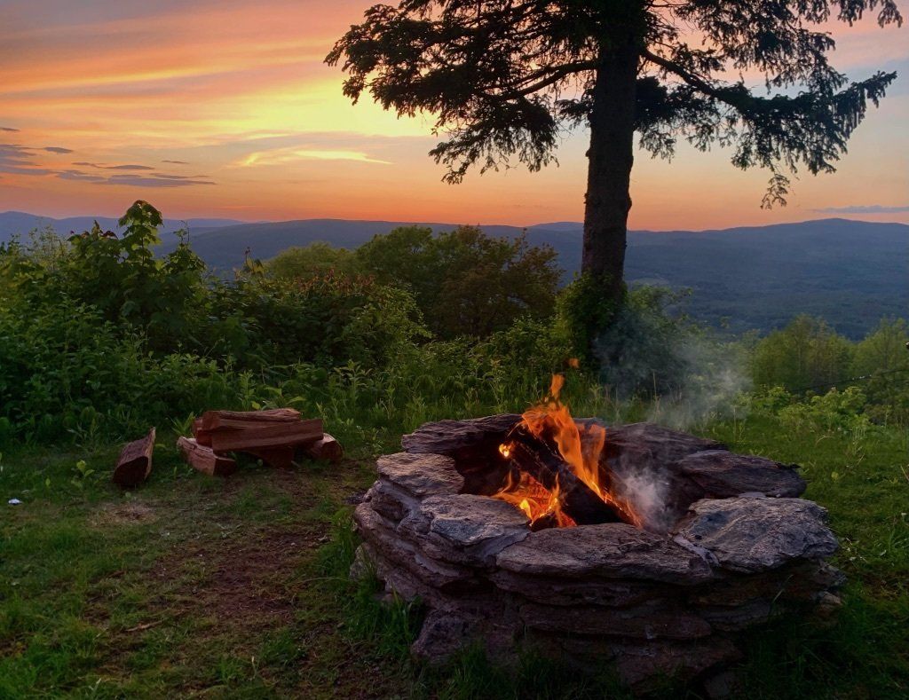 A stone fire pit with a crackling fire at dusk, set on a grassy hill overlooking a mountain range under a sunset sky.