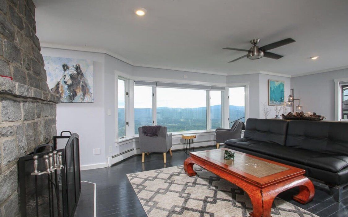 A living room with a stone fireplace, black leather sofa, patterned rug, and a scenic view of mountains through windows.