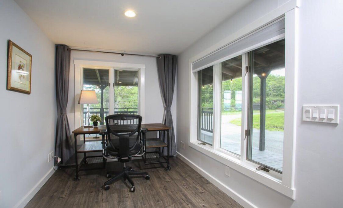 A bright home office with a desk, black mesh chair, and large windows looking out onto a grassy yard.