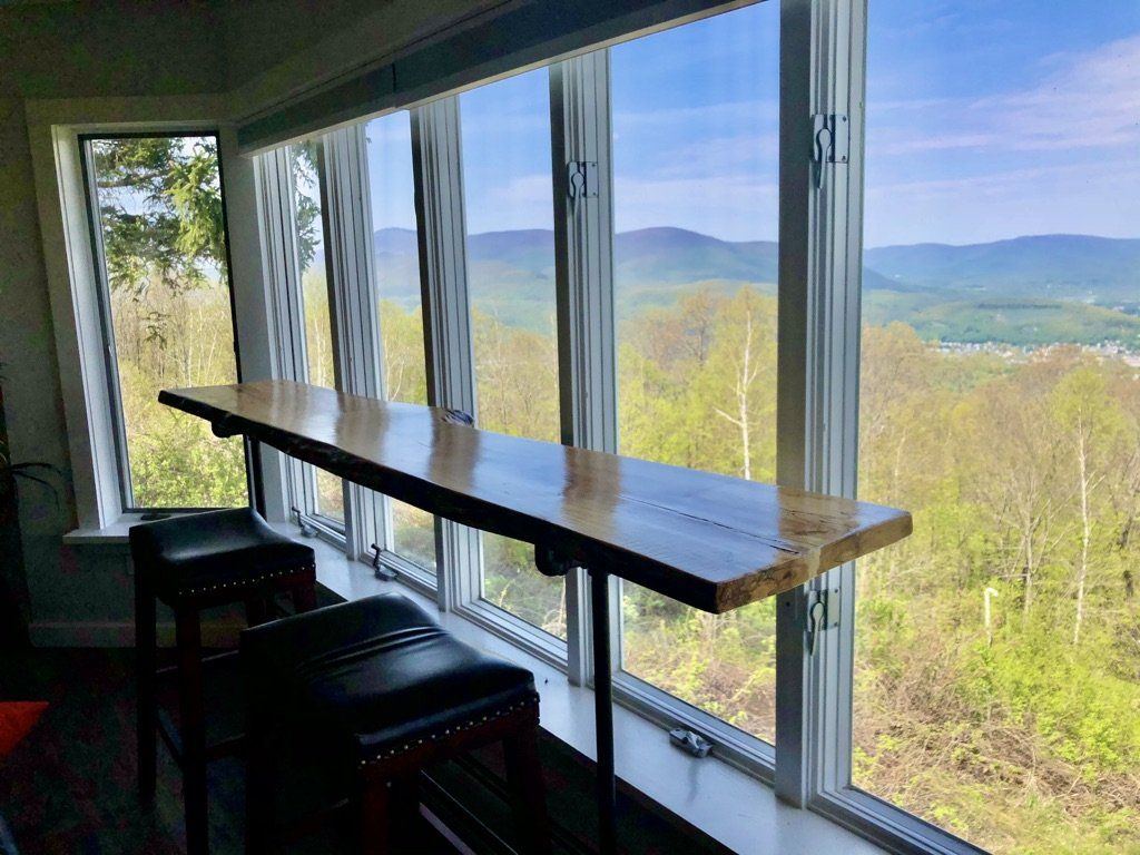 A natural wood bar top sits in front of a floor-to-ceiling window overlooking a mountain range, with two stools nearby.