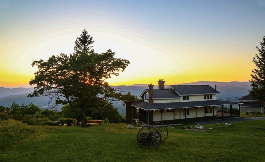 A white house with a porch sits on a grassy hill at sunset, with a large tree in the foreground and mountains beyond.