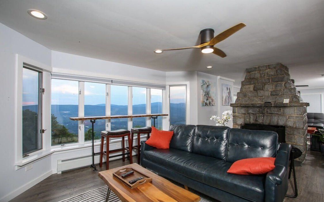 A cozy living room with a leather sofa, orange pillows, a stone fireplace, and a window bar overlooking a mountain view.