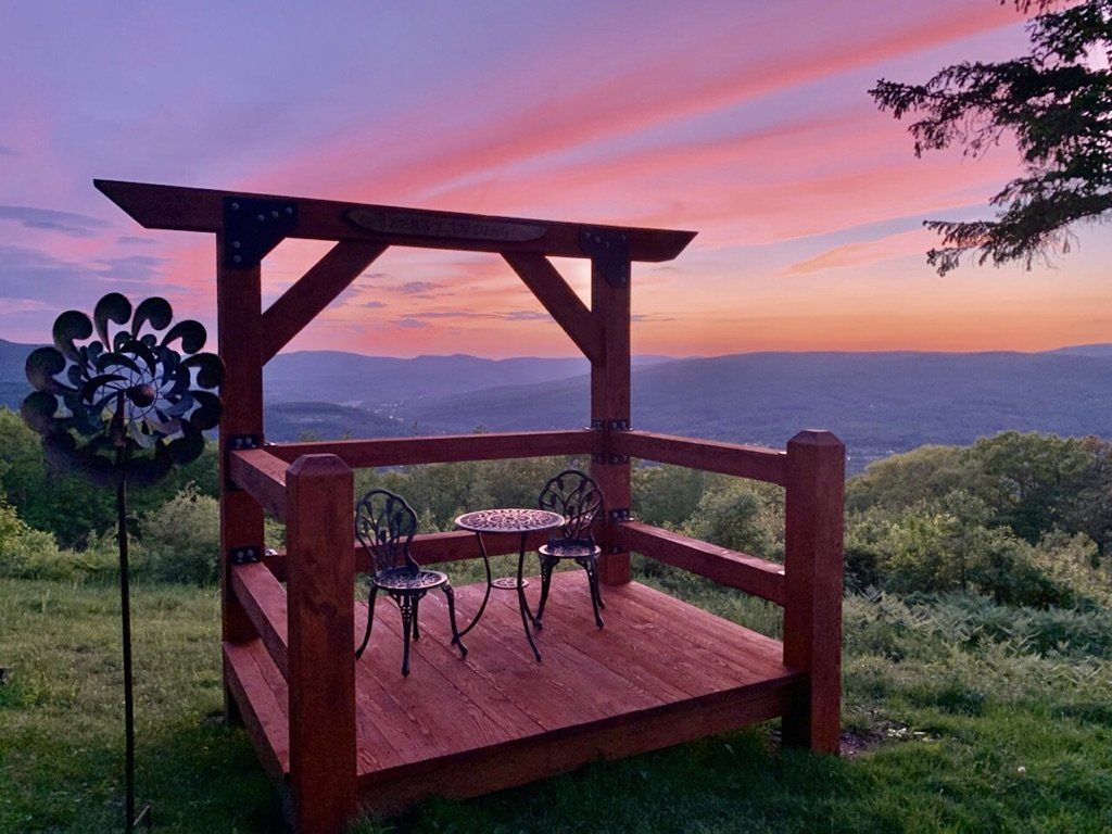 A wooden deck with a table and two chairs overlooks a scenic valley under a vibrant, colorful sunset sky.