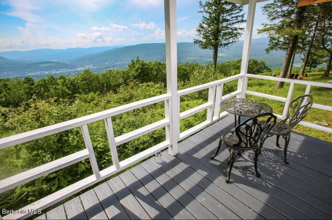 A view from a wooden deck with black metal chairs and a table, overlooking a lush green valley and mountains.