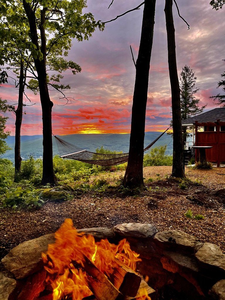A fire pit burns in the foreground with a hammock and cabin set against a vibrant, orange sunset over rolling hills.