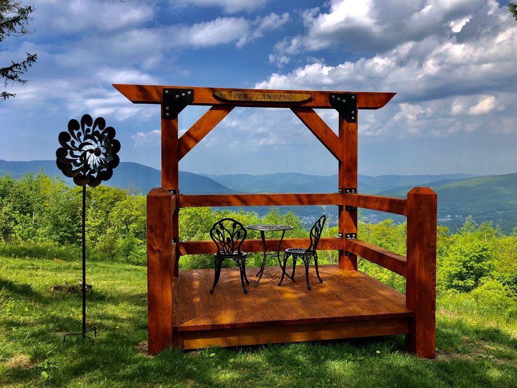 A wooden gazebo frame sits on a grassy hill overlooking a scenic mountain valley, with a small table and two chairs inside.