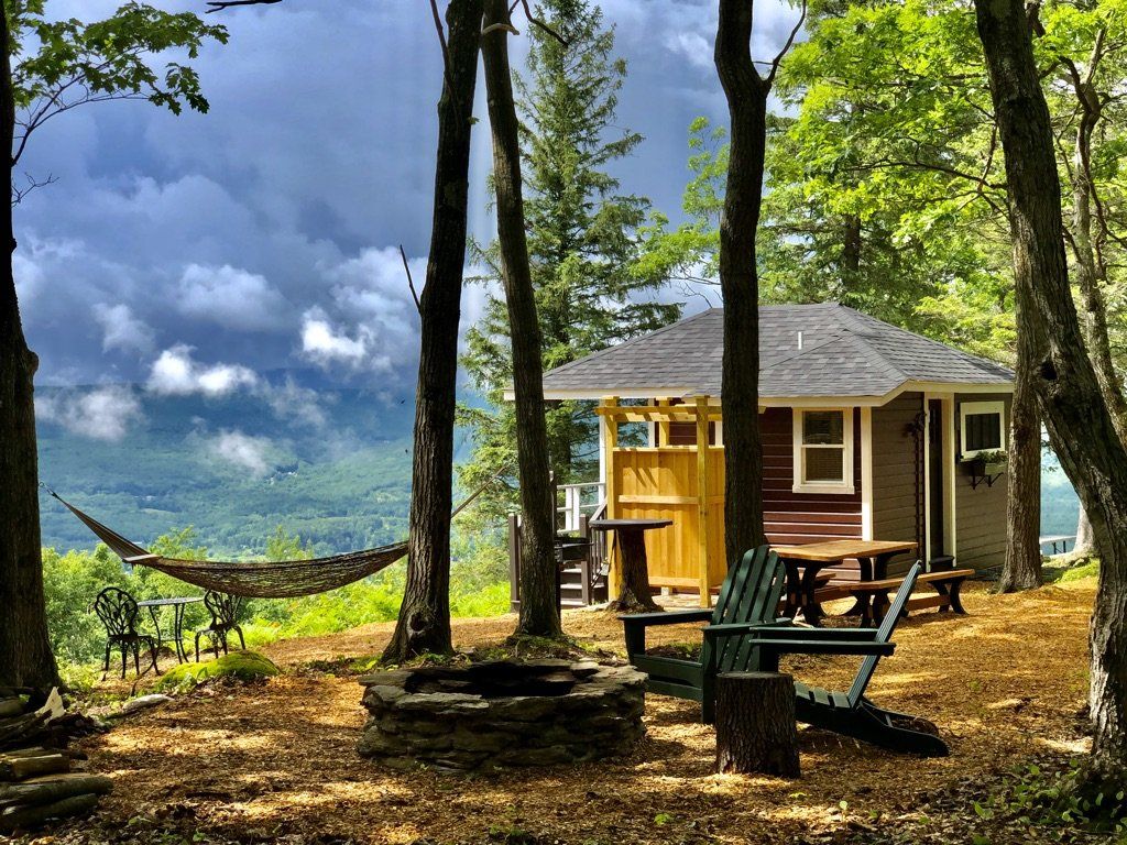 A cabin with a fire pit, hammock, and Adirondack chairs sits in a wooded clearing overlooking a mountain valley.