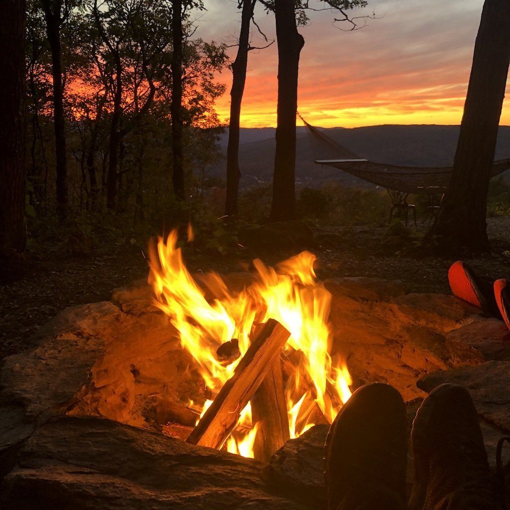 A campfire burns in a stone pit at dusk, with a hammock silhouetted against a vibrant orange and purple sunset.