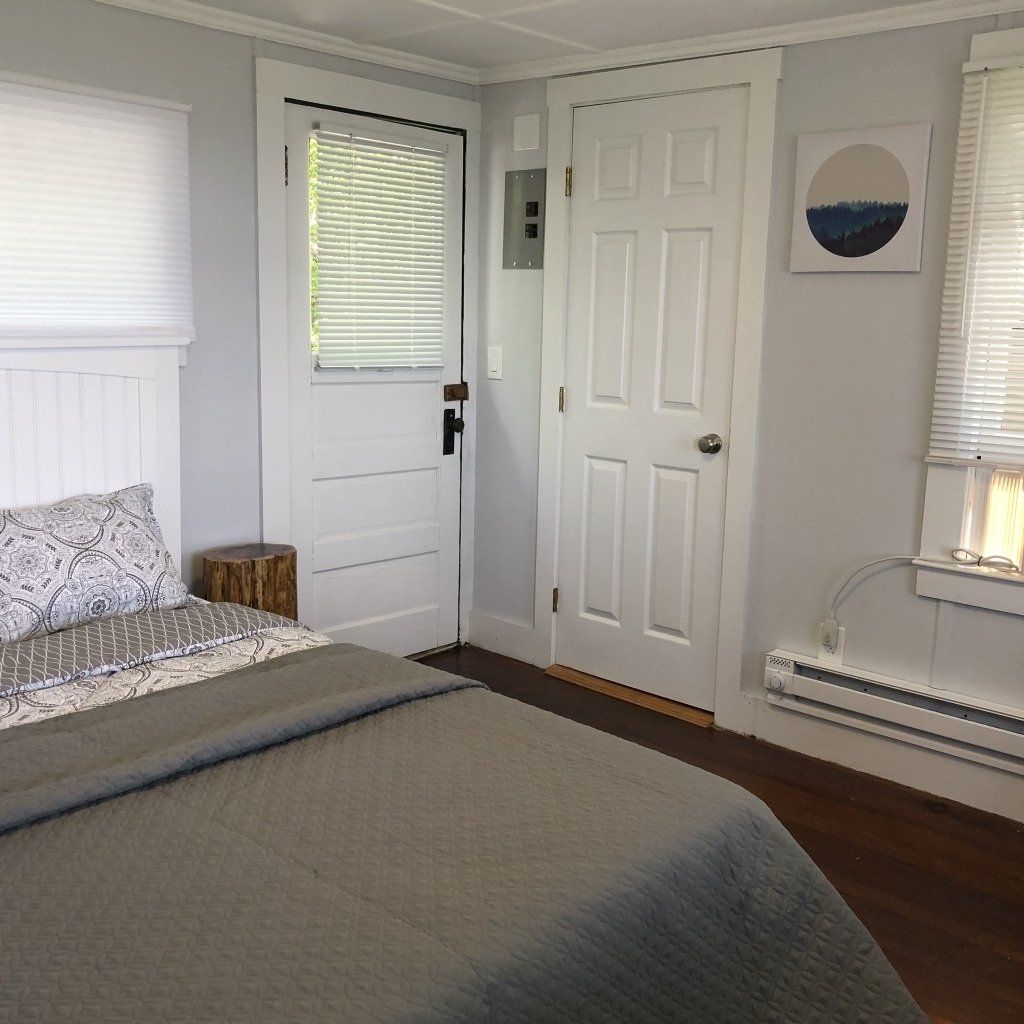 A bedroom featuring a bed with a gray quilt, a white door, a glass-paned door with blinds, and a piece of circle art.