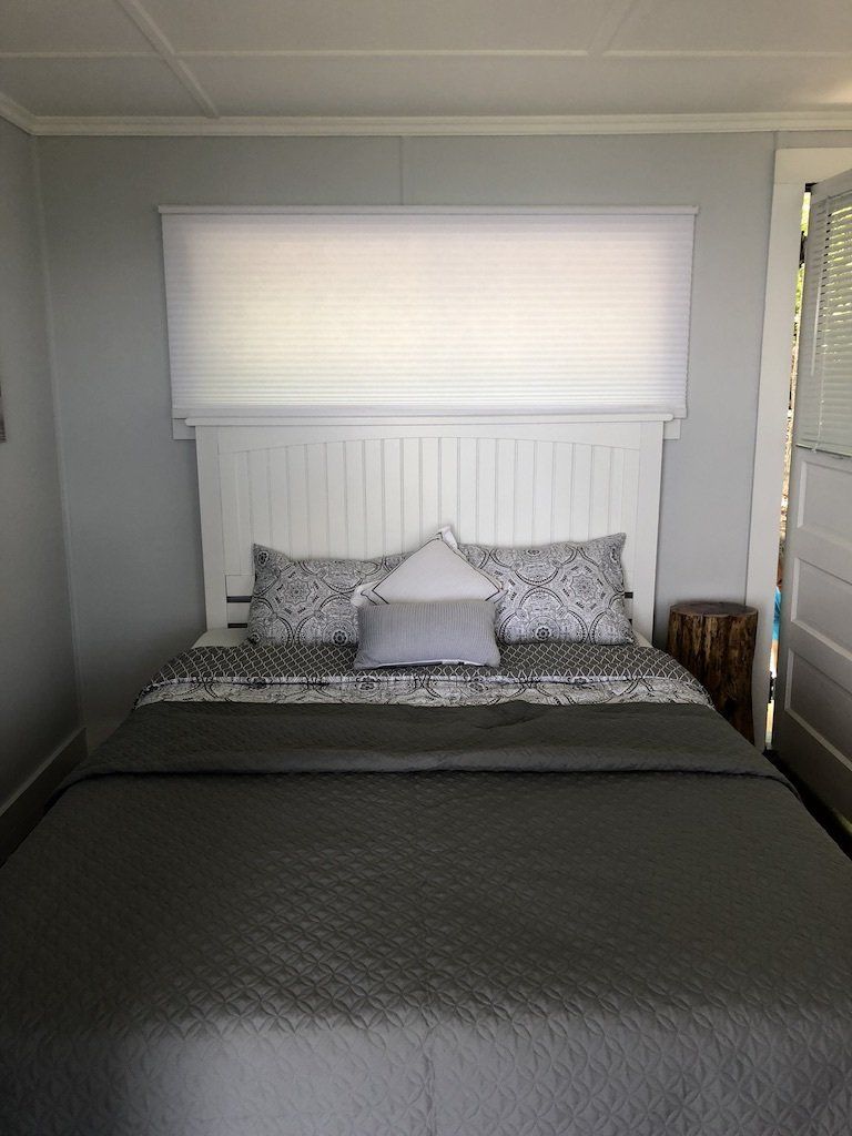 A neatly made bed with a dark gray quilt, patterned pillows, and a white paneled headboard under a wide window blind.