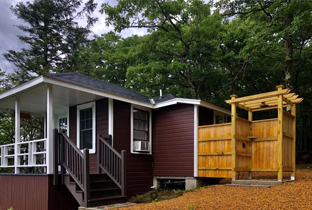 A small dark brown cottage with a white-railed porch and a wooden outdoor shower enclosure set in a wooded area.