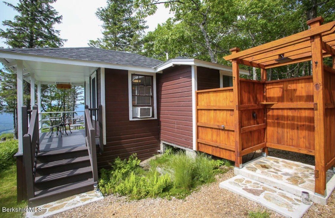 A dark brown cabin with a porch and steps leads to an outdoor wooden shower stall set in a lush, green woodland area.