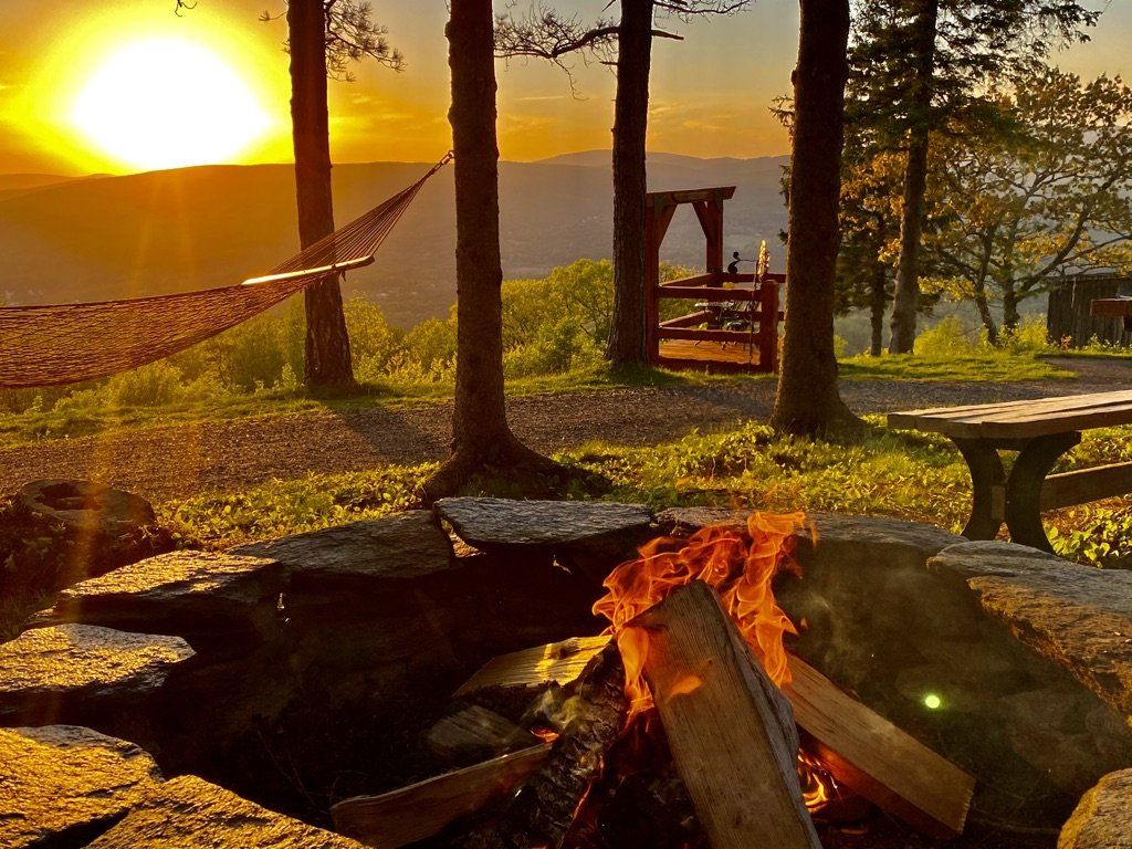 A stone fire pit with a crackling fire at sunset, overlooking a scenic valley with a hammock and trees.