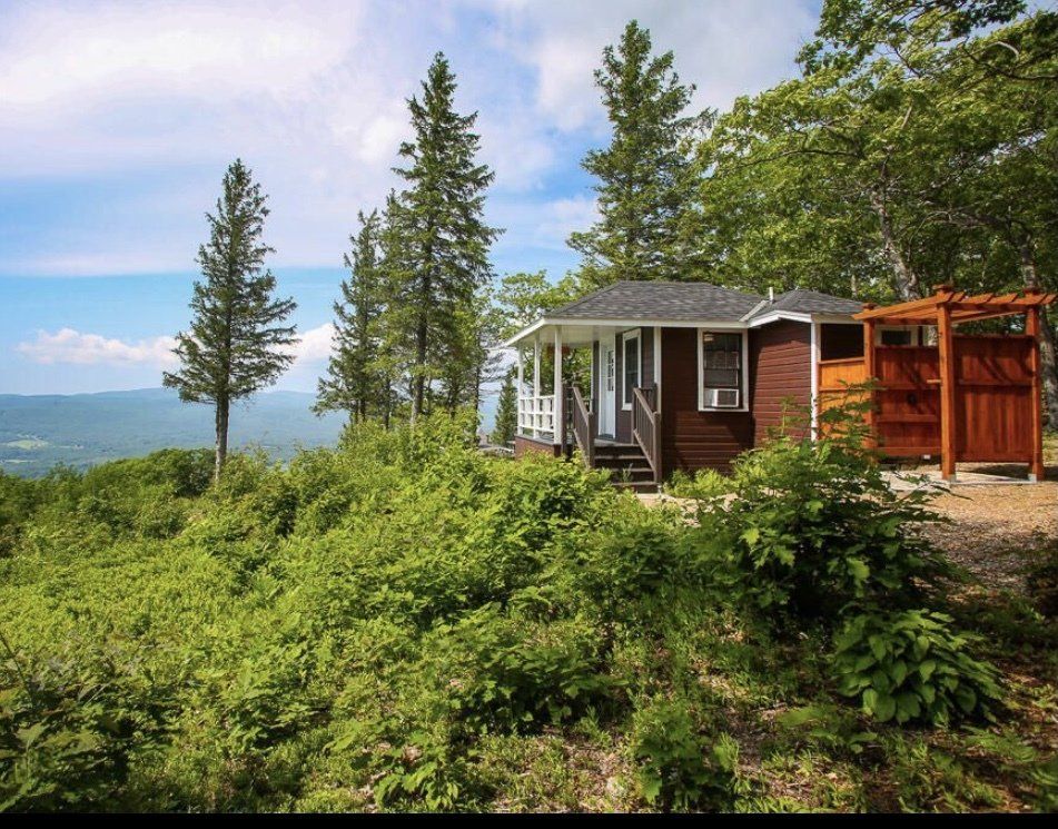 A small, brown cabin with a porch and wooden pergola sits on a hillside overlooking a vast landscape of trees and mountains.