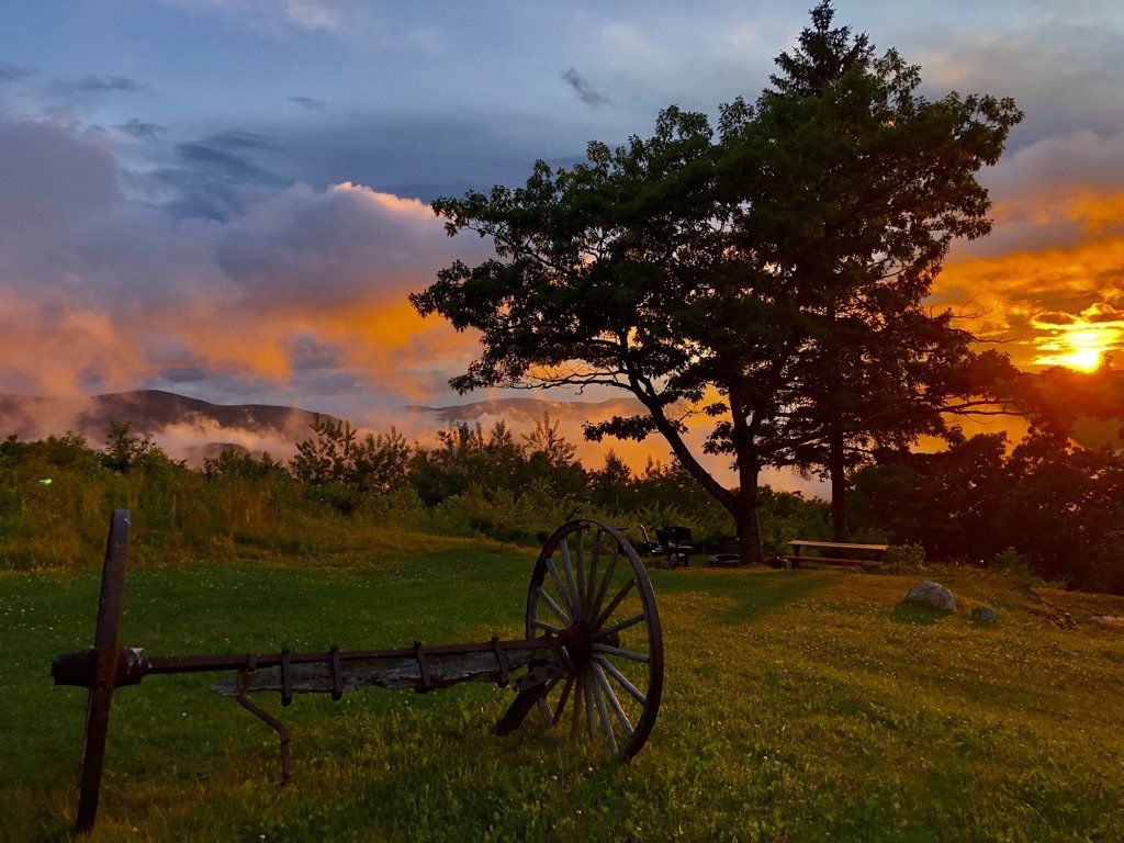 A vintage farm implement sits in a grassy meadow under a dramatic sunset, with a large tree silhouetted against the sky.