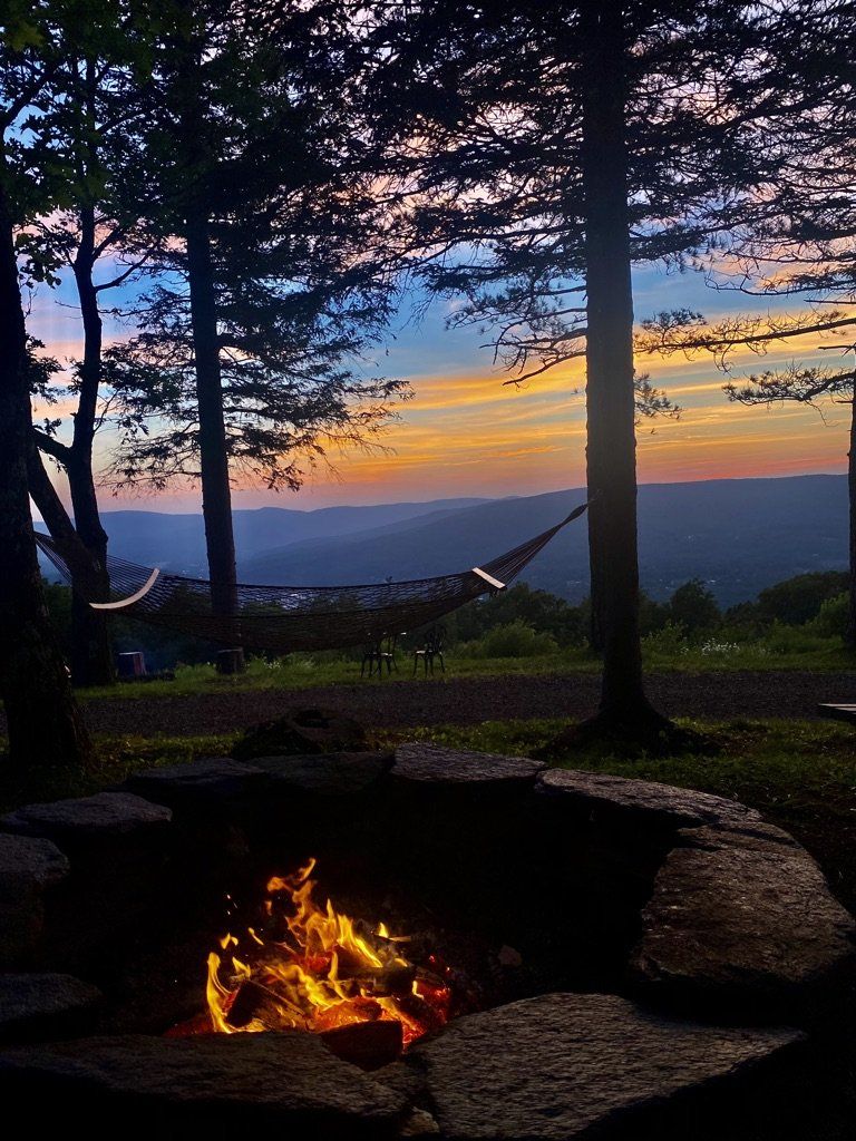 A campfire burns in a stone ring at dusk, with a hammock hanging between trees overlooking a mountain valley sunset.