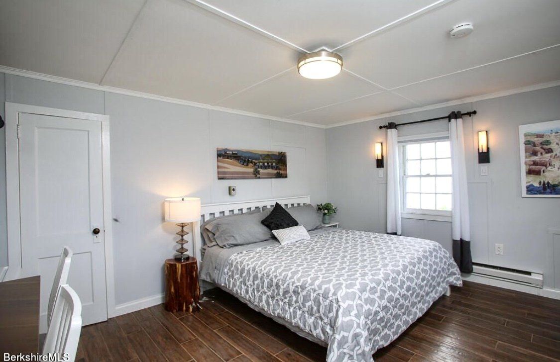 A bright bedroom featuring a queen bed with a patterned comforter, light gray walls, wooden floors, and two wall sconces.