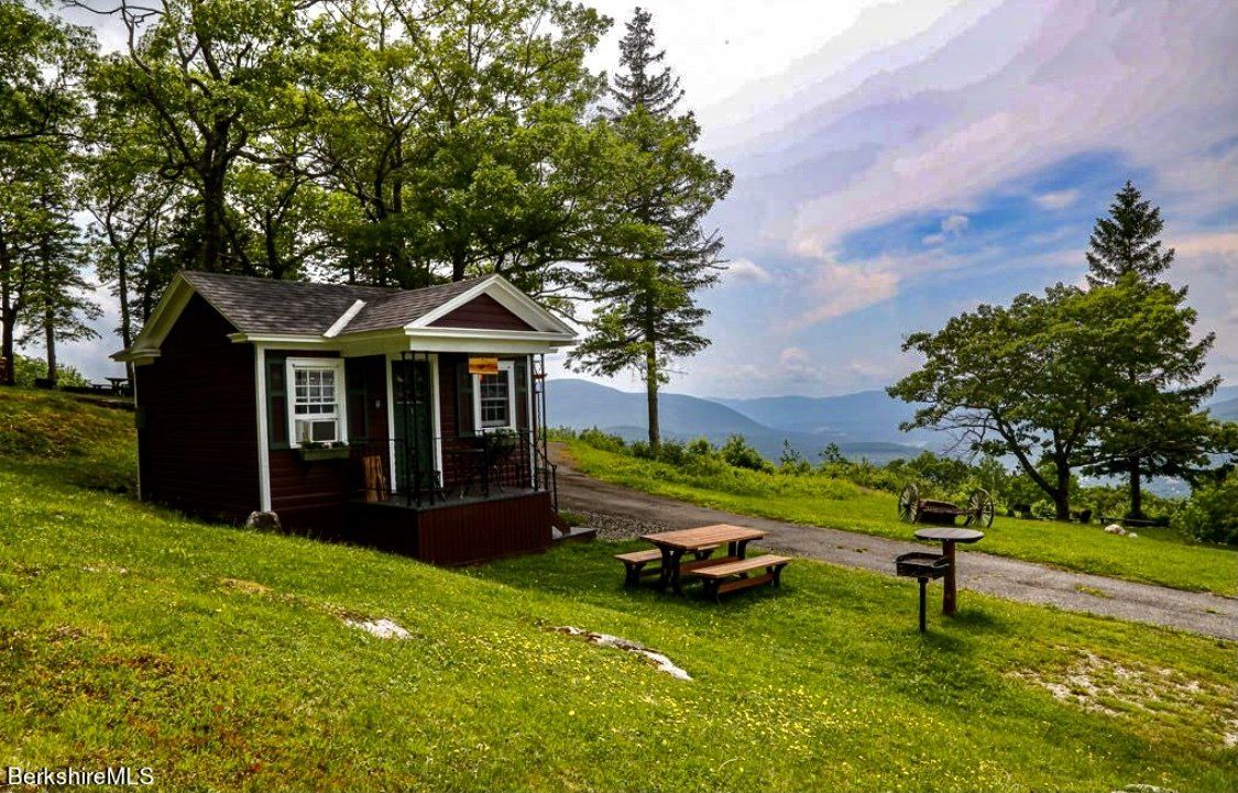A small, dark red cabin with a picnic table sits on a grassy hill overlooking a mountain range under a cloudy sky.