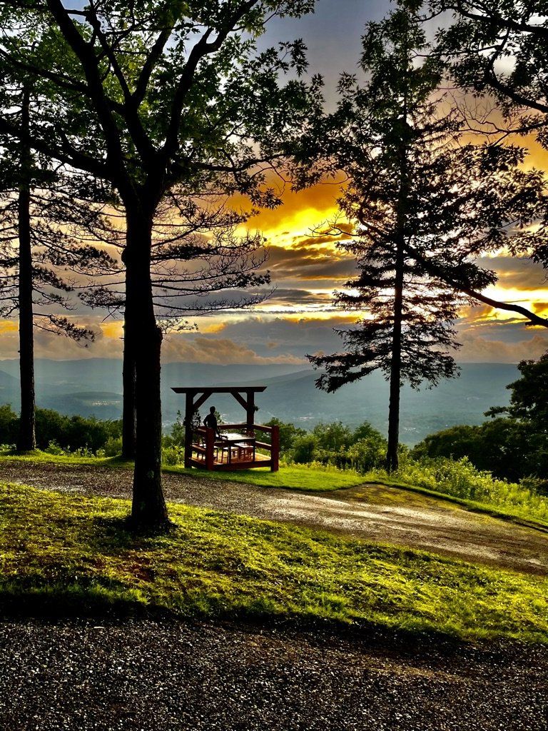 A wooden picnic shelter overlooks a scenic mountain valley under a vibrant, golden sunset sky framed by silhouetted trees.