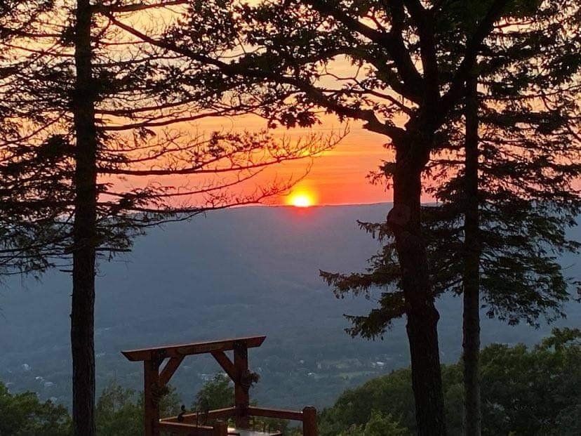 A vibrant sunset glows behind silhouetted trees overlooking a distant valley with a wooden structure in the foreground.
