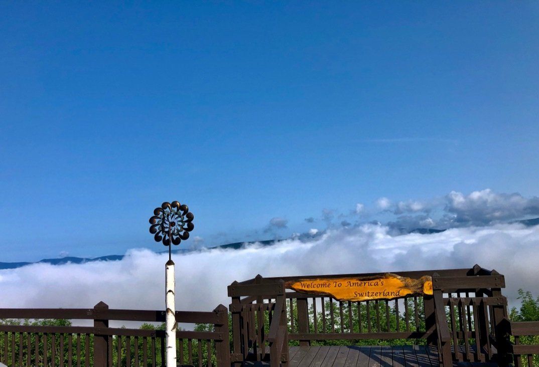 A wooden observation deck overlooks a thick sea of white clouds under a clear blue sky, with a decorative metal spinner.