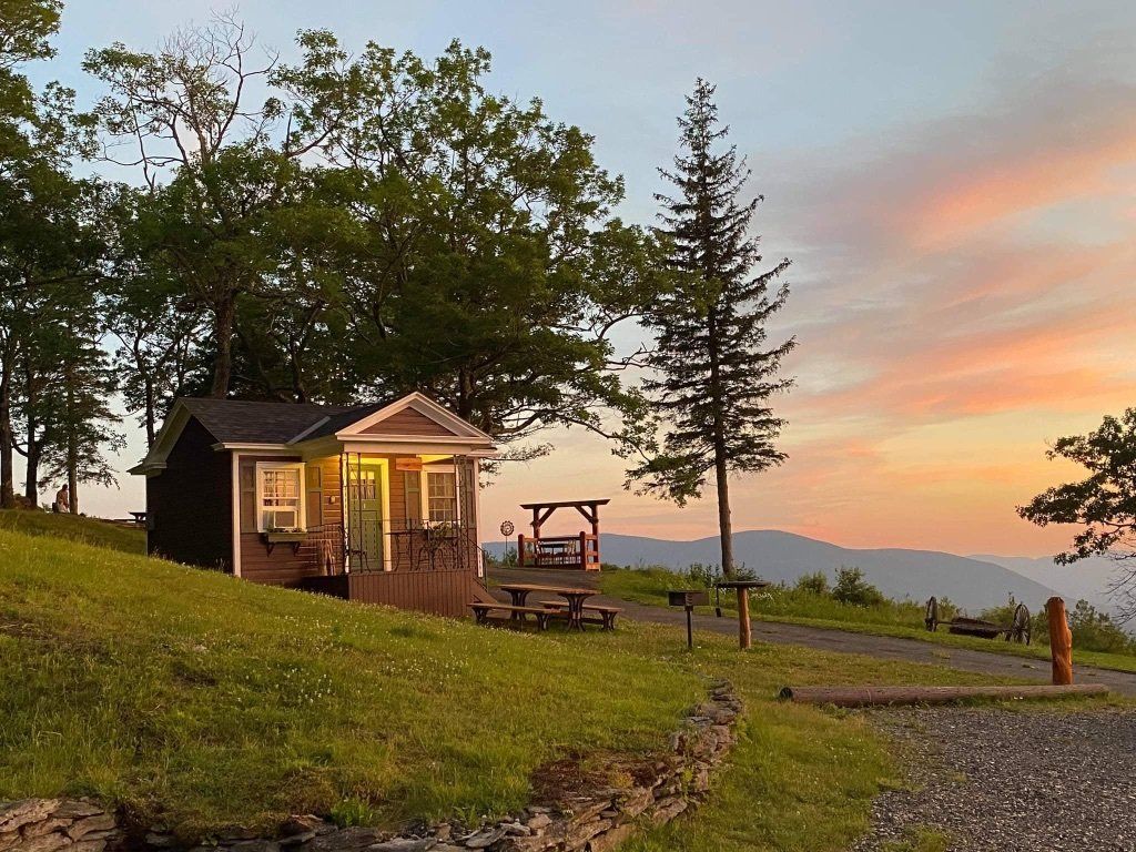 A small, brown cottage sits on a grassy hill at sunset, overlooking distant mountains under a pastel sky.