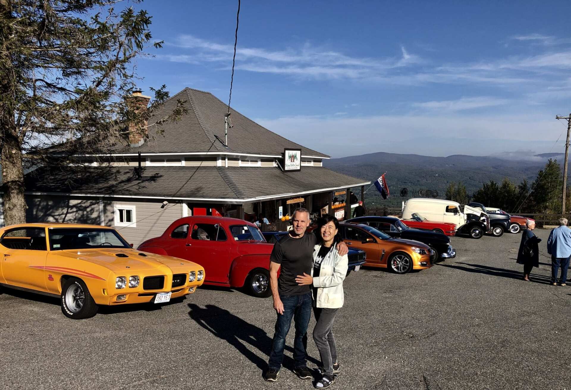 A couple poses in a gravel lot filled with classic cars in front of a rustic mountain building under a sunny sky.
