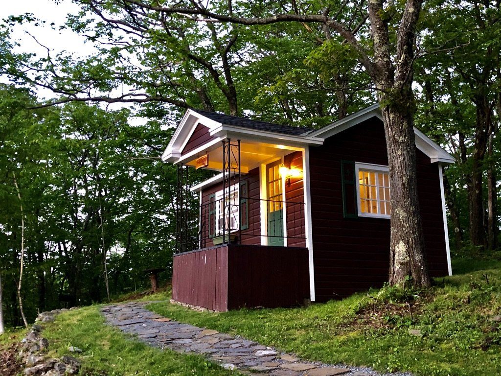 A small, dark red cabin with a lighted porch and a stone path, nestled among lush green trees in a wooded setting.