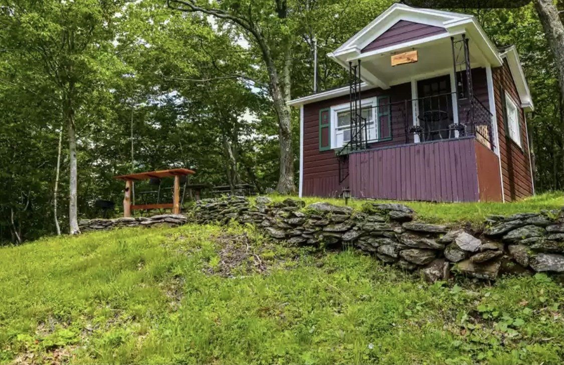 A small, purple wooden cabin with a porch sits on a grassy hill behind a stone retaining wall in a forest.