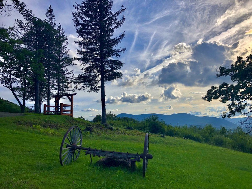 An old farm rake sits on a grassy hill overlooking a mountain range under a cloudy sky at sunset.