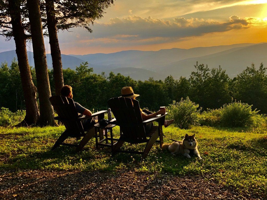 Two people sit in chairs on a grassy hill at sunset, watching a mountain view with a dog resting nearby.