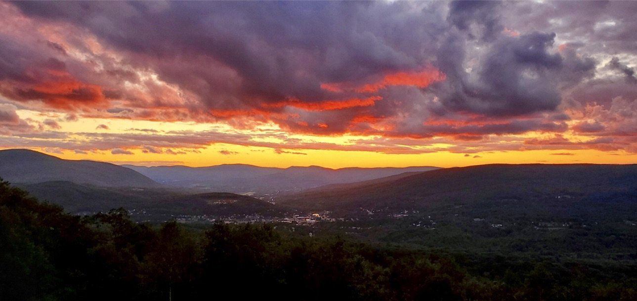 A vibrant sunset with orange, pink, and dark clouds over a forested mountain valley with a small town in the distance.