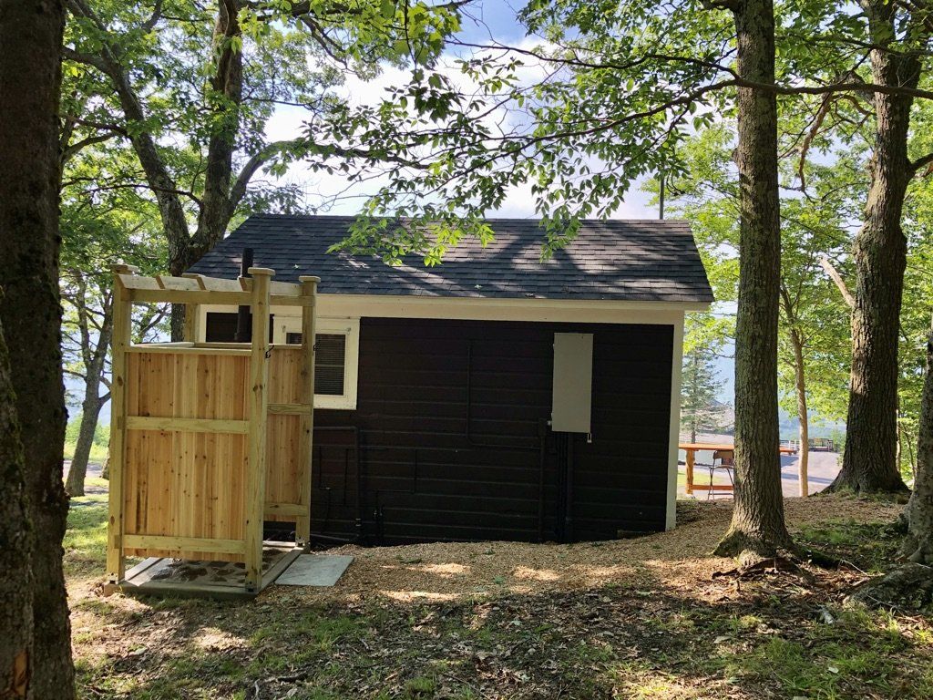 A dark-sided small shed set in a wooded area with an attached wooden outdoor shower enclosure.