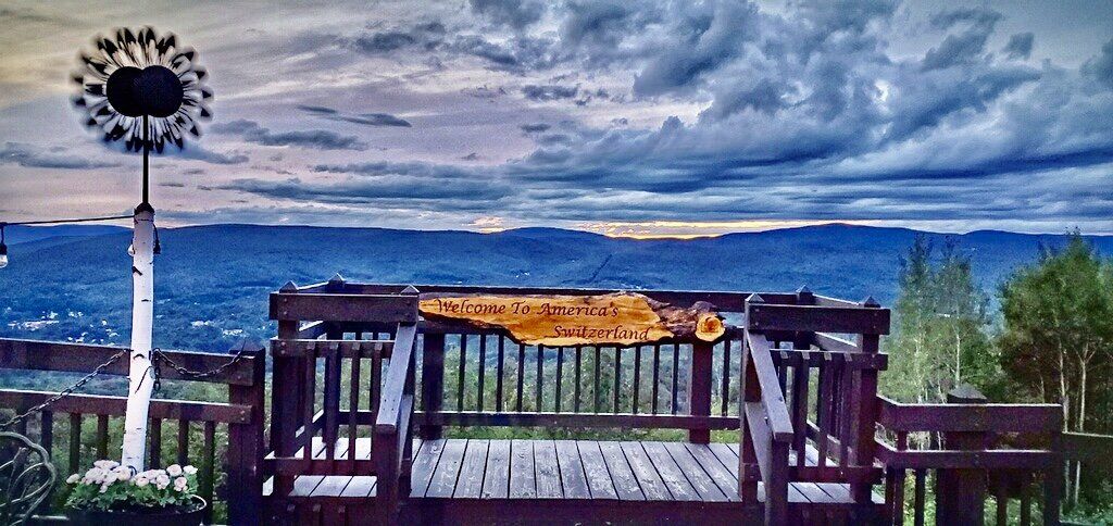 Wooden deck viewing platform with a metal windmill, overlooking a deep blue mountain valley under a cloudy sunset sky.