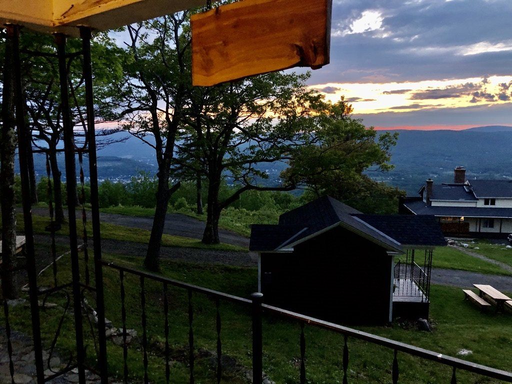 View from a porch overlooking a hillside with trees, a small dark shed, and a sunset over a distant mountain valley.
